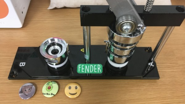 Fender badge maker with three badges - a cat badge, an avocado badge, and a smiley face badge.