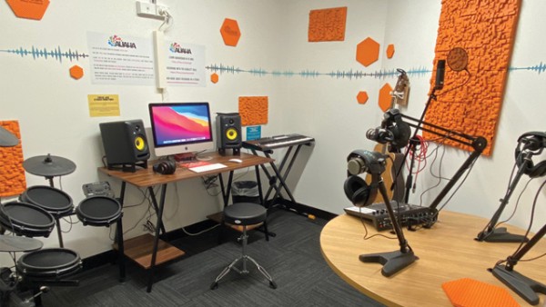 Recording studio with iMac and speakers at a desk, with keyboard and electronic drums on either side, and another table with microphone stands and headphones. There are different shaped orange acoustic panels on the white walls.
