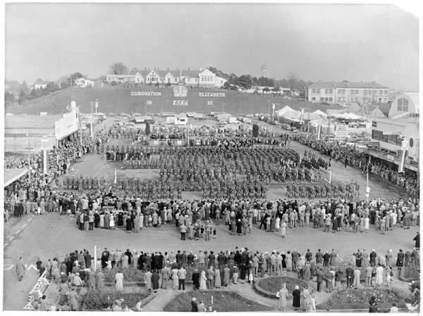 A black and white image showing a crowd gathered in an open square. The centre of the square features a military parade.