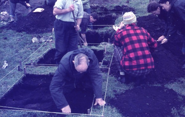 A colour image showing seven people on an archaeological dig. Three men are standing inside squares that are being excavated, the other four people are crouched next to the open holes putting dirt through a sieve. 