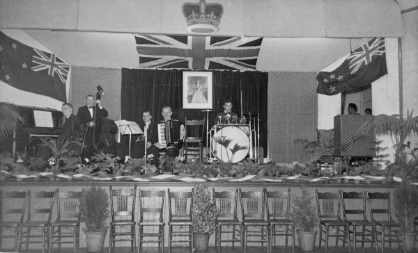 A black and white image showing a band on a stage. The stage is decorated with plants, two New Zealand flags and a Union Jack. A portrait of Queen Elizabeth II hands behind the band. The band consists of a pianist, double bassist, trombonist, accordionist and drummer.