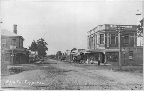 Old dirt street with horse and carts. Wooden buildings with verandahs on left, brick building with verandahs on right.