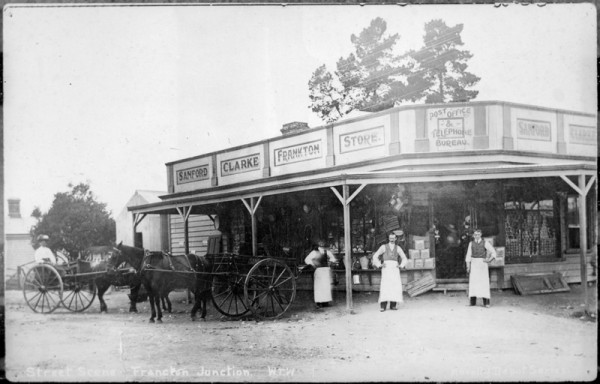 Image of old wooden store. Men in long aprons standing out front, alongside two horse and carts.