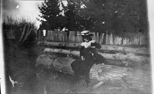 A black and white image showing a girl dressed in a long black dress with white collar and straw hat, sitting on a log with a black and white cat next to her.