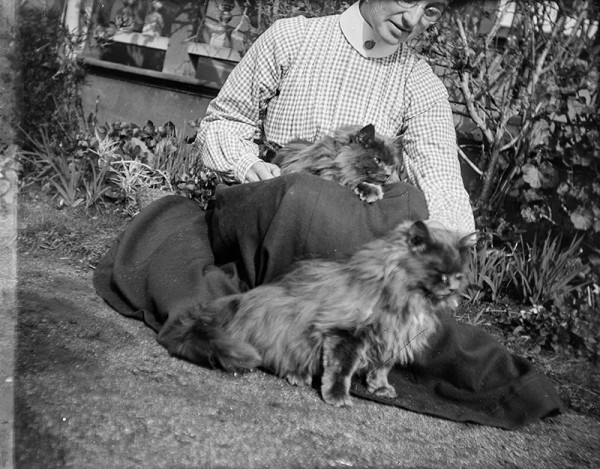 A black and white image showing a woman dressed in a long dark skirt and checked long sleeve shirt, seated on grass next to a garden, holding a dark fluffy cat on her lap. A second dark fluffy cat sits on her skirt.
