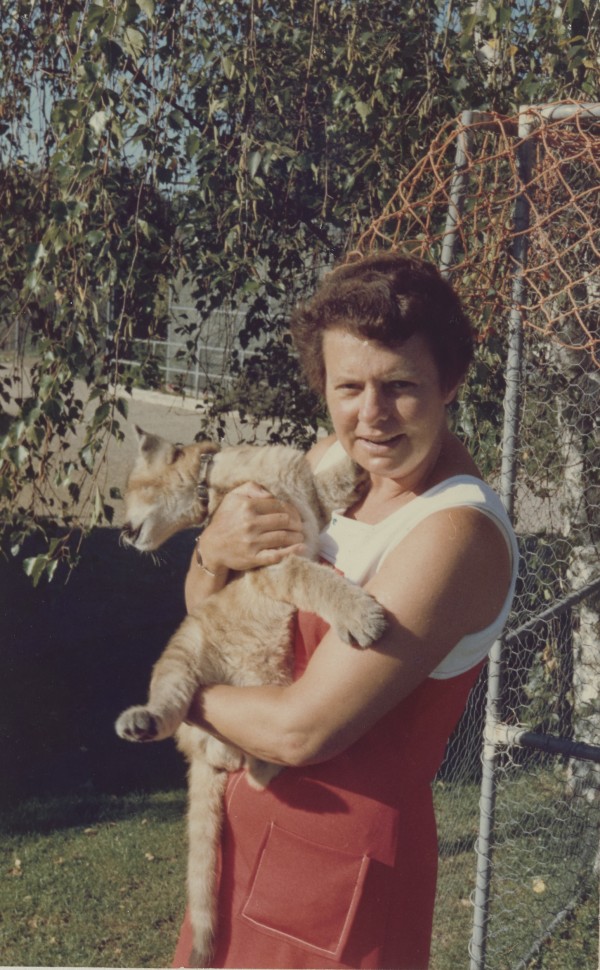 A colour image showing a woman in a red and white dress holding a lion cub in her arms. The cub is twisted away from the camera.