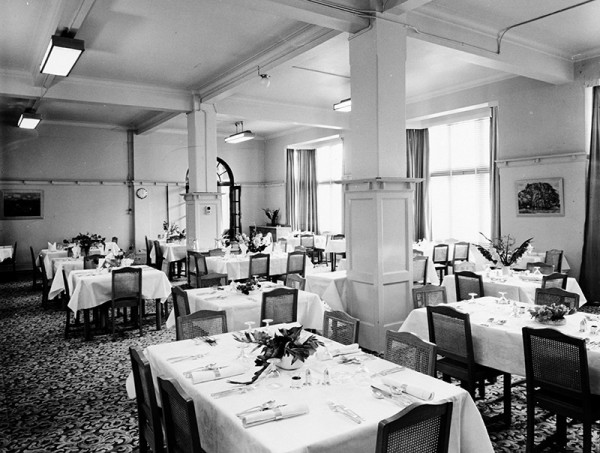 A black and white image showing a hotel dining room with at least ten tables laid with cutlery, napkins and centrepieces.