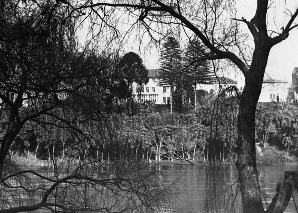 A black and white image showing a river scene bordered by trees. A large white hotel is visible on the opposite riverbank through the trees.