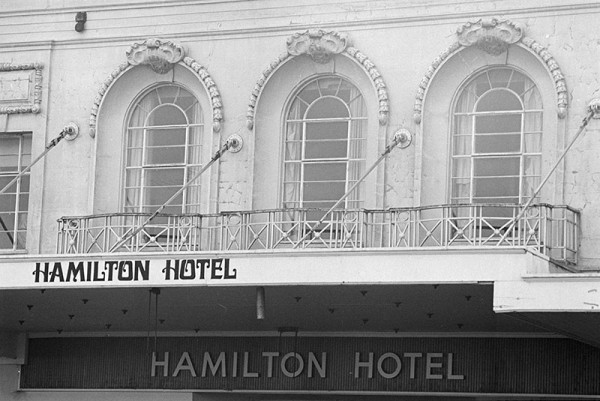 A black and white image showing the balcony of the Hamilton Hotel. Three large arched windows look out to a metal balcony.