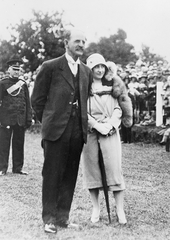 A black and white image showing  woman and man in 1930s style dress standing on grass. In the background of the image there is a man in military uniform and a large crowd. The main subjects of the image are Mayor J F Fow and the Duchess of York.