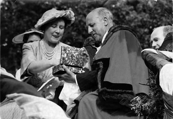 A black and white image showing a man in mayoral robes offering a gift to a woman in pearls and a hat seated next to him.