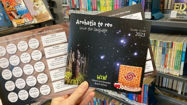 Programme booklets held up in front of a bookshelf. One booklet is closed, showing the front page with Matariki in the sky, and a second booklet behind it is open, showing the reading log to be filled out.