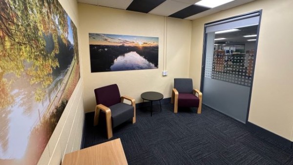 Interior of the Kirikiriroa Room. The walls are a soft yellow colour, with peaceful images of nature hanging. There are two chairs and a small side tables at one end of the space, and a window out into the library with a blind that can be closed.