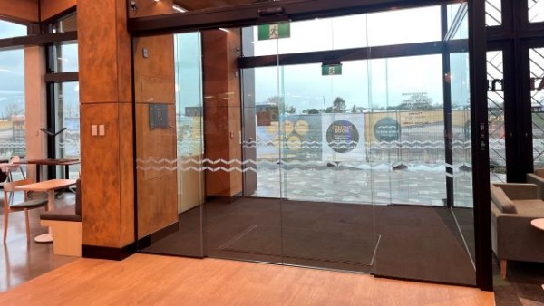 South entrance of Te Kete Aronui from the inside looking out. There are two sets of double-wide automatic doors leading to a paved pathway.