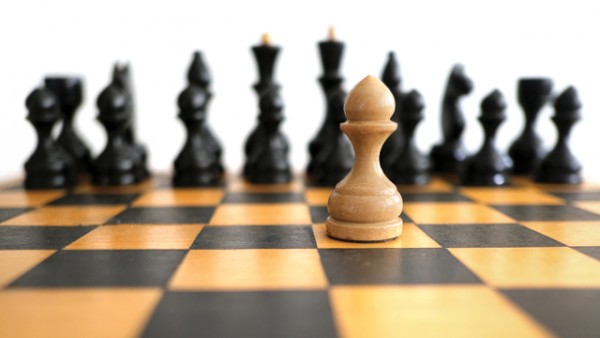 A wooden chess board with black wooden chess pieces in the background, out of focus, and a light-coloured wooden pawn piece in the foreground, in focus.