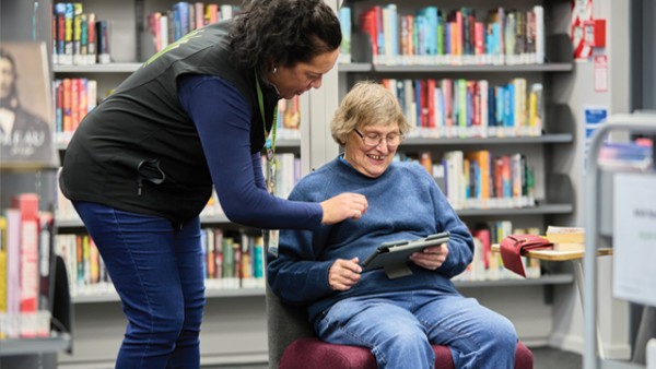 A librarian, standing, assisting an older person, who is sitting, with using a tablet.