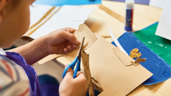 Over-shoulder shot of a child using scissors to cut shapes from cardboard. There is other craft material on the table in front, like cardboard, coloured paper, and a glue stick.