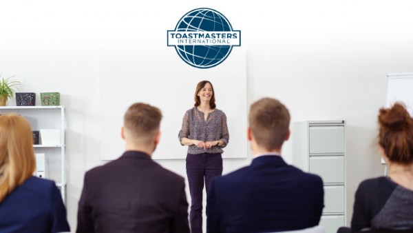 Toastmasters logo above woman standing to speak in front of an audience.